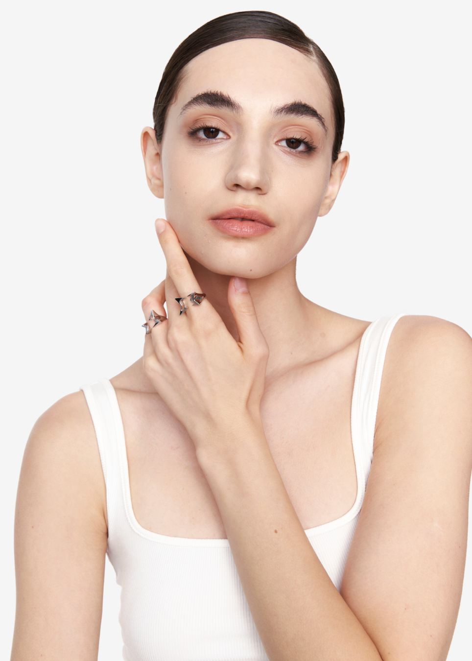 A close-up portrait of a model wearing multiple silver rings on her fingers, dressed in a white tank top against a clean white background.