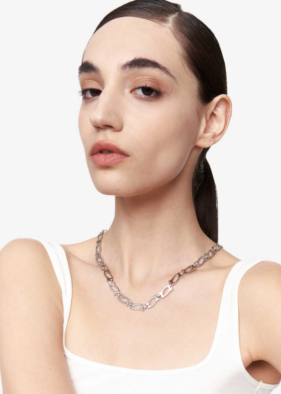 A close-up of a model wearing a silver-toned chain necklace with a heart-shaped link design, paired with a white tank top against a light gray background.