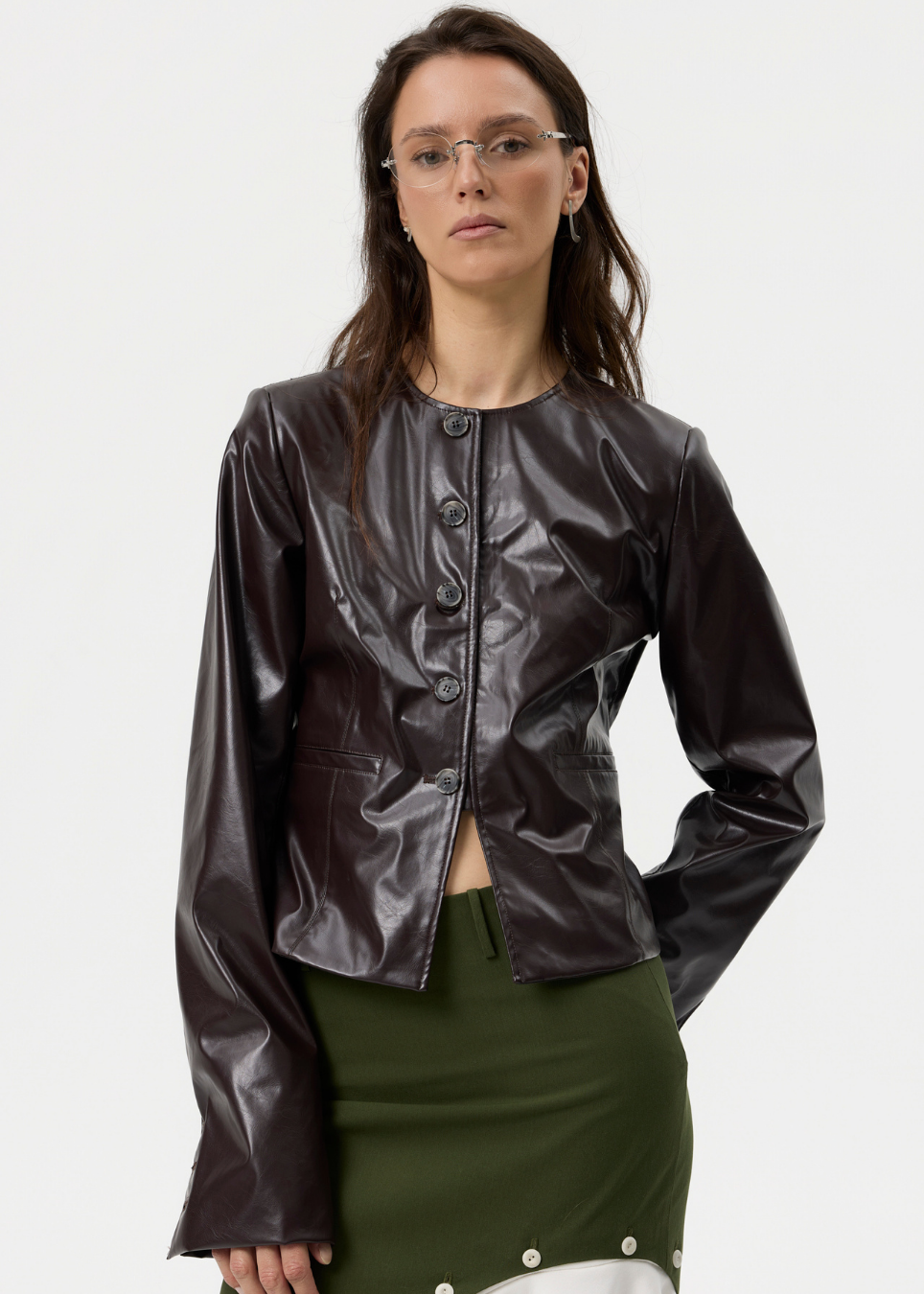 A model wearing a cropped, long-sleeve dark brown faux-leather jacket with a button front, paired with an olive green skirt, against a plain white studio background.
