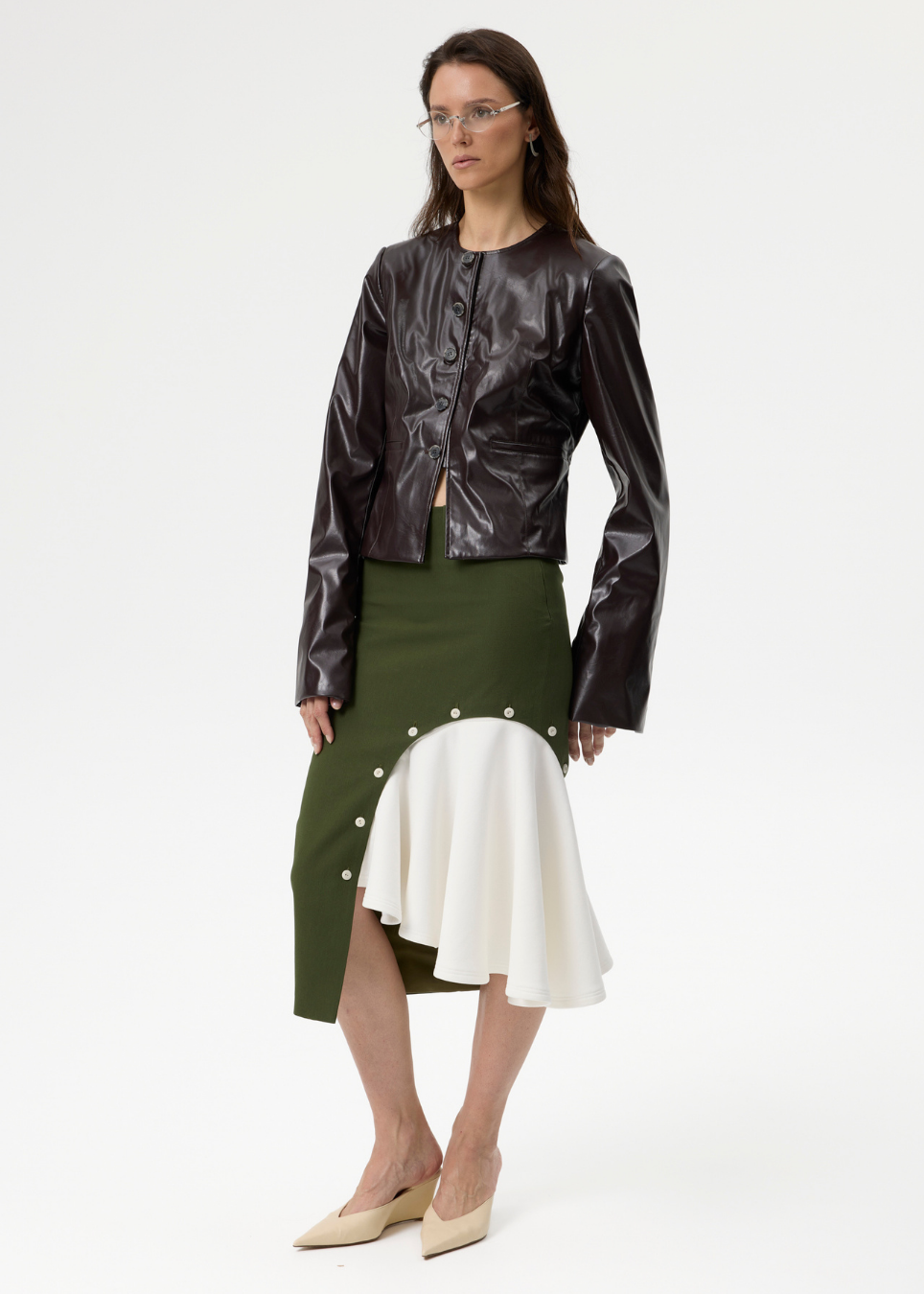 A model stands against a plain white studio background wearing a dark brown glossy cropped jacket, an olive green skirt with a white ruffled overlay and decorative buttons, and beige pointed-toe heels.