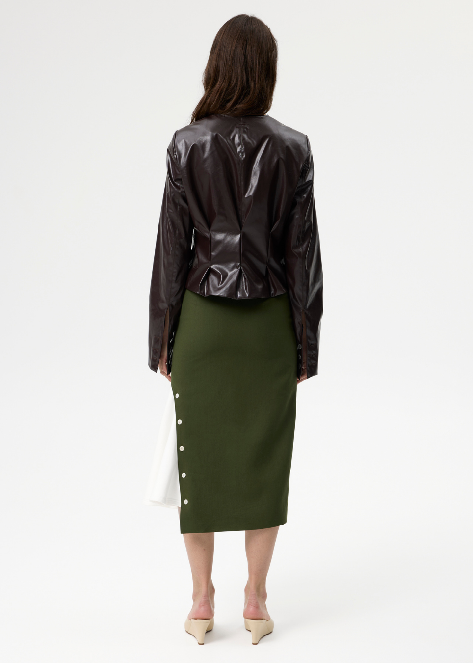 A model shown from the back wearing a dark brown glossy cropped jacket and an olive green knee-length skirt with white buttons down the side, paired with beige heels against a plain white studio background.