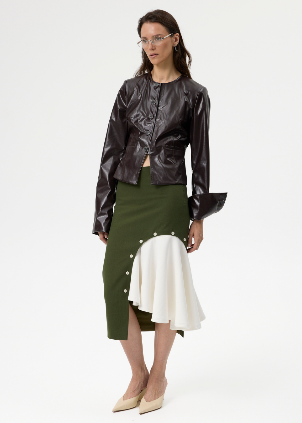 A model stands against a plain white studio background wearing a dark brown glossy cropped jacket, an olive green and white asymmetrical skirt with pearl buttons, and beige pointed-toe heels.