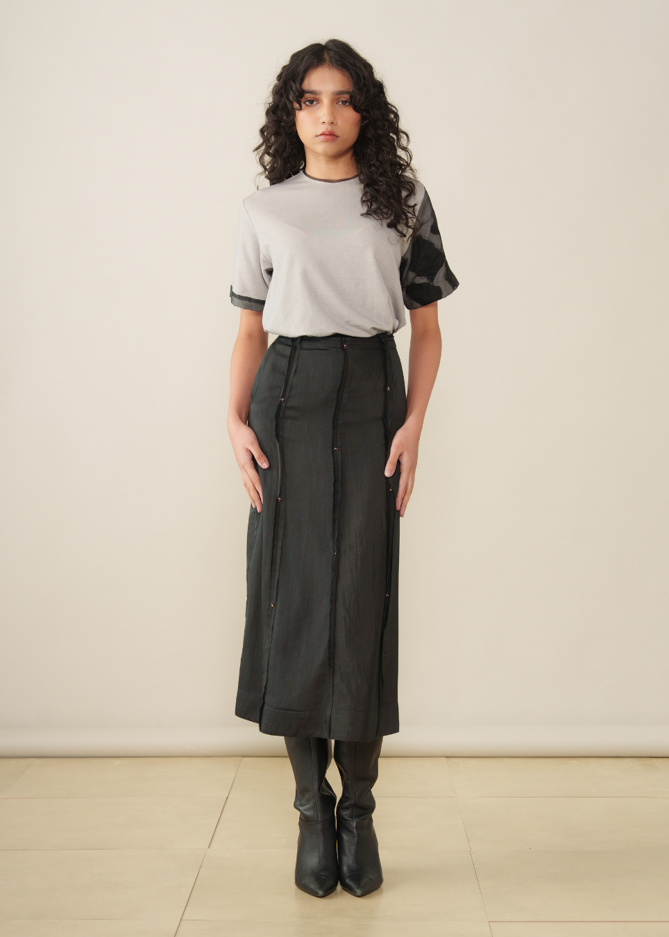A model stands in a studio wearing a light gray short-sleeve top with black sheer flutter sleeves, paired with a black midi skirt, black knee-high socks, and black ankle boots.