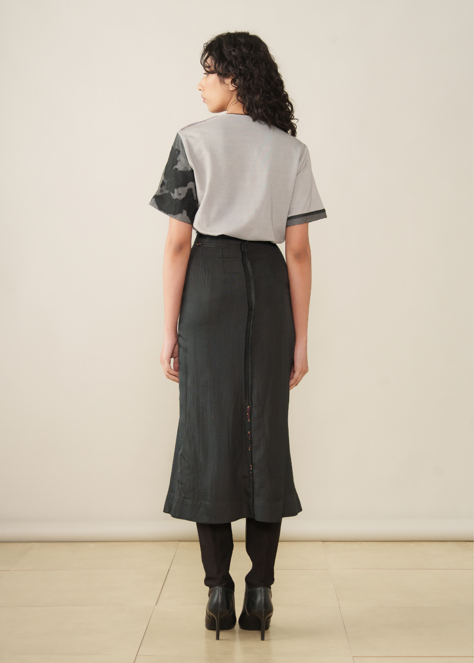 A model is shown from the back wearing a short-sleeve top with a light gray body and dark patterned sleeves, paired with a dark gray pleated midi skirt, black tights, and black ankle boots, set against a plain beige studio background.
