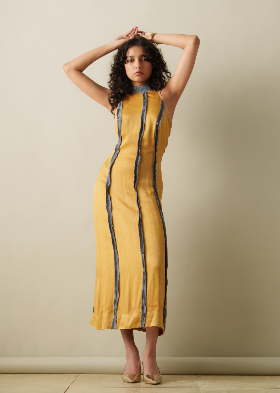 A woman modeling a sleeveless mustard yellow midi dress with vertical dark blue stripes, standing against a plain beige wall.