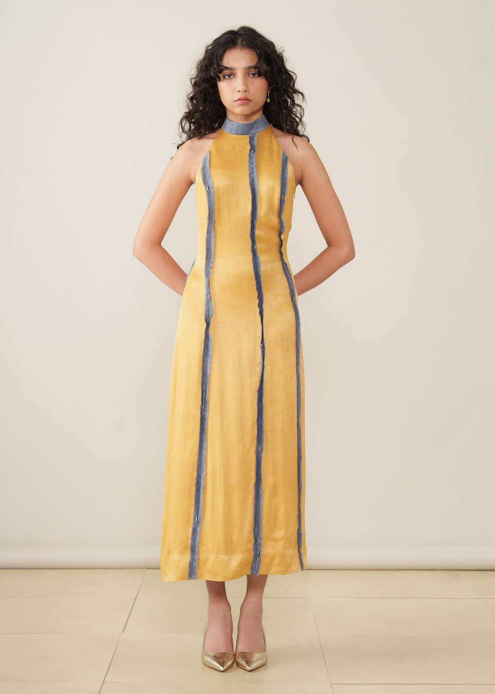 A woman modeling a sleeveless yellow midi dress with vertical blue stripes and a high blue collar, standing against a plain beige wall.