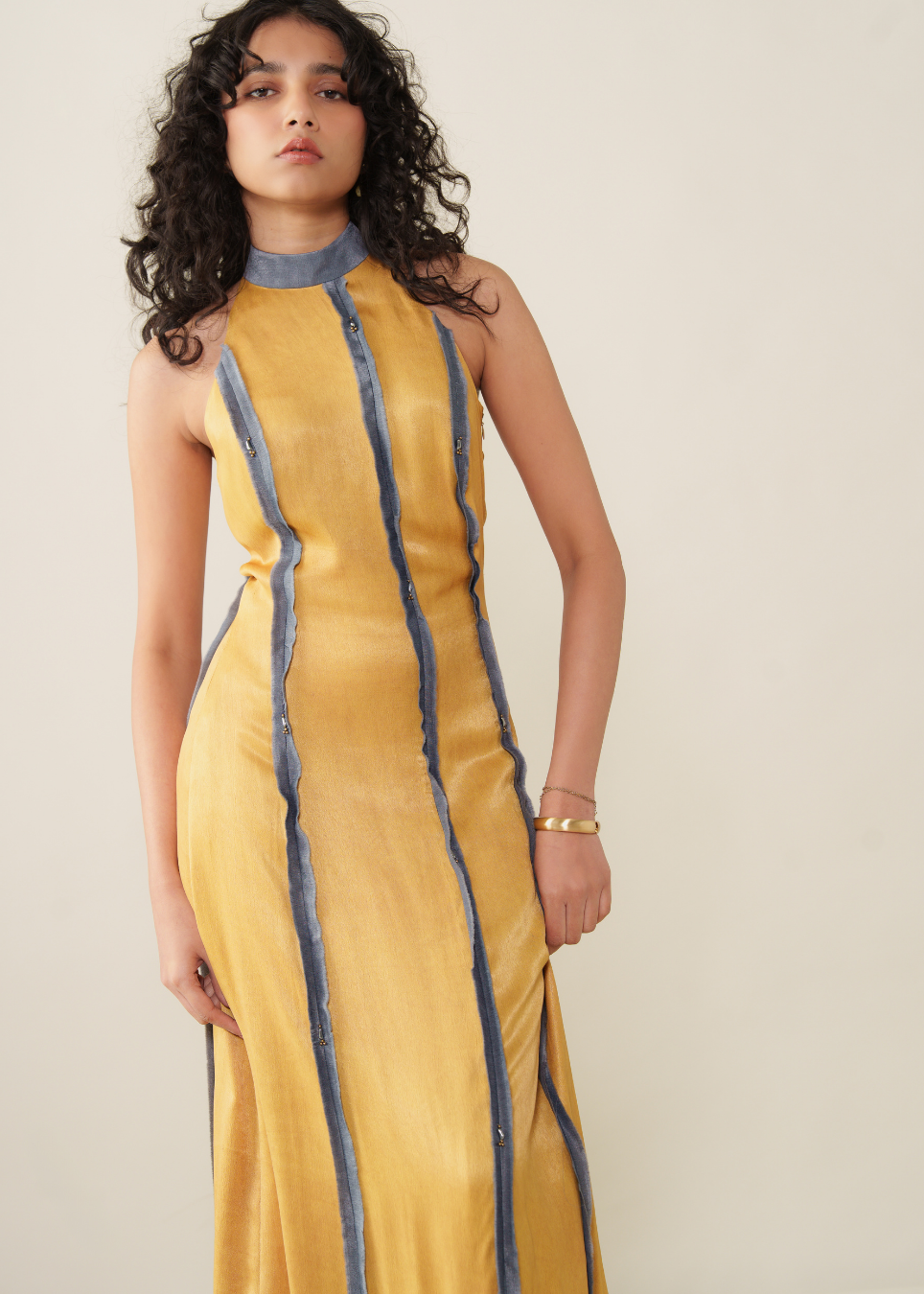A woman modeling a sleeveless mustard yellow maxi dress with vertical blue-gray stripe details and a matching mock neck, set against a plain beige studio background.