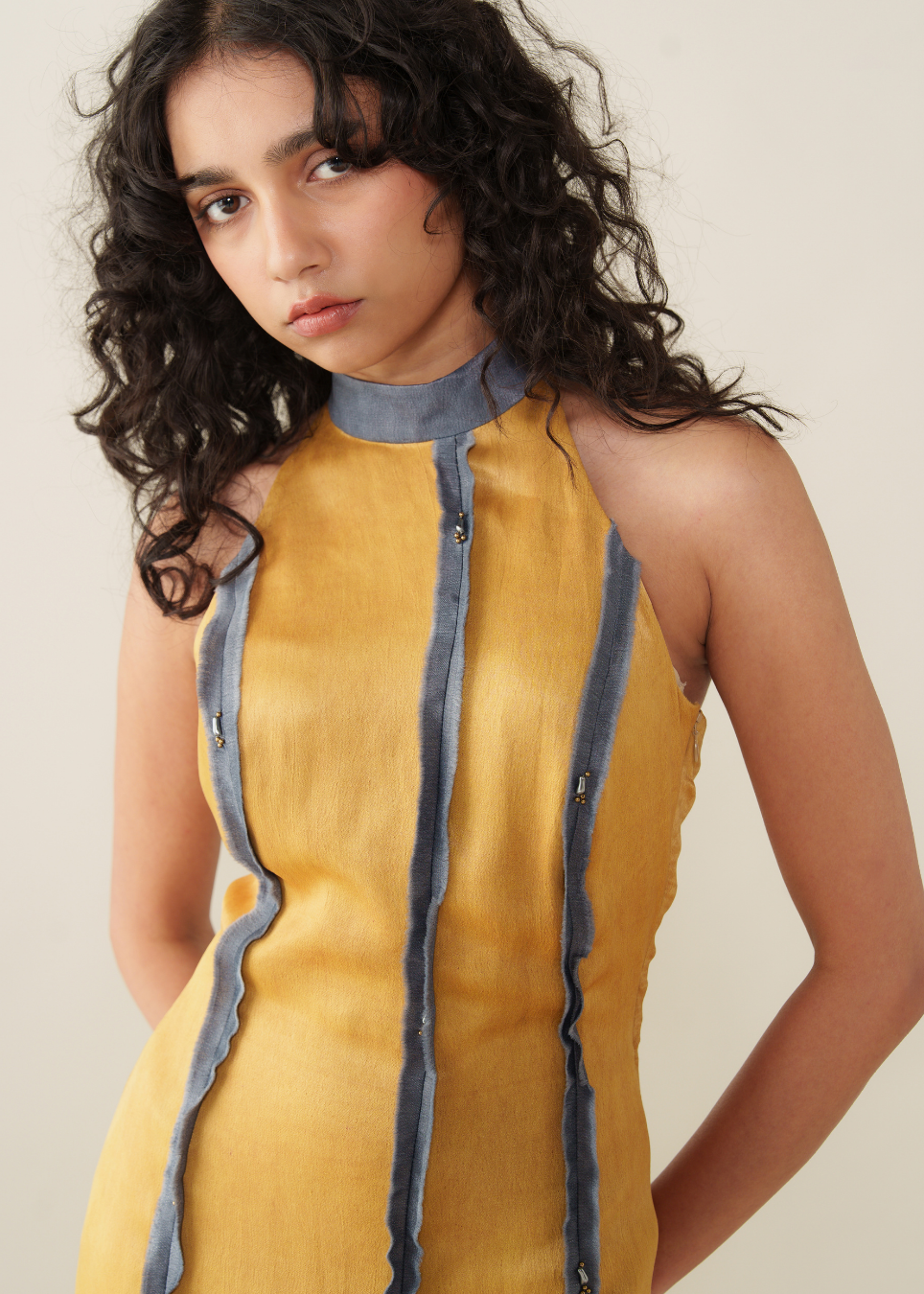 A sleeveless mustard yellow dress with a gray mock neck and vertical gray trim details, modeled by a woman with curly dark hair against a plain beige background.