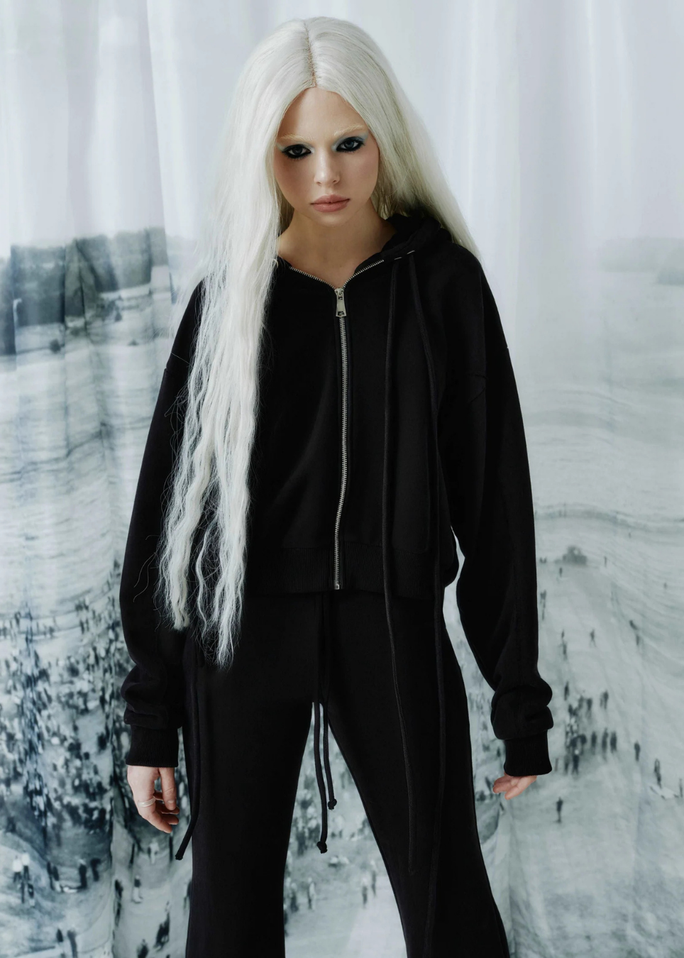 A model with long platinum blonde hair wearing a black zip-up hoodie and matching black sweatpants, standing in front of a white curtain backdrop with a black and white landscape print.