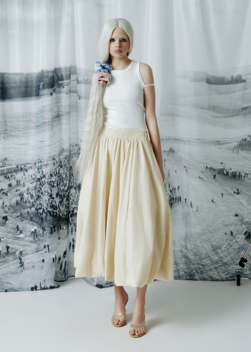A model with long platinum blonde hair stands in front of a large black and white scenic backdrop, wearing a white cold-shoulder top and a sand-colored pleated maxi skirt.