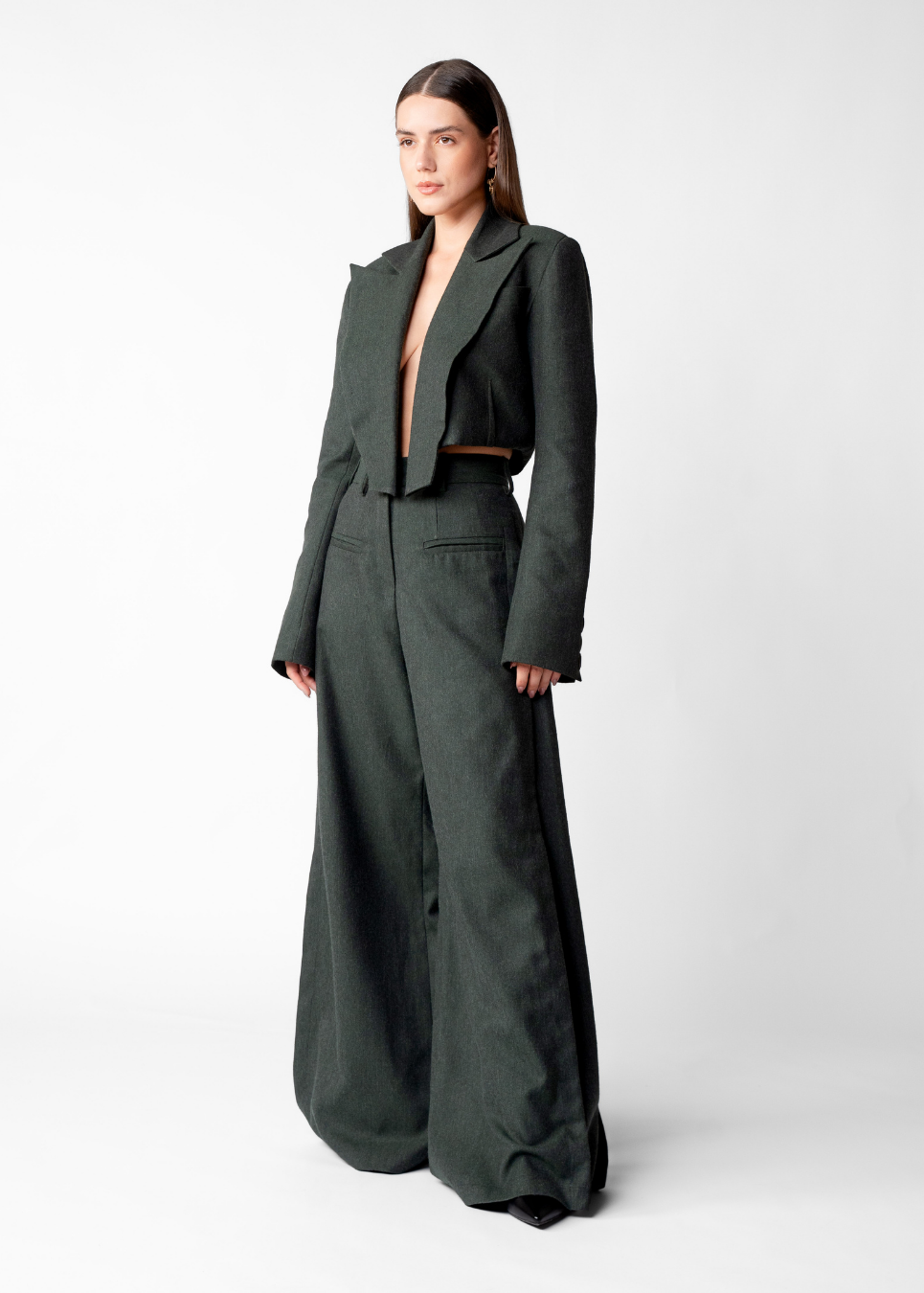 A model stands against a light gray studio background wearing a dark olive green two-piece outfit consisting of a cropped blazer-style jacket with wide lapels and matching wide-leg trousers.