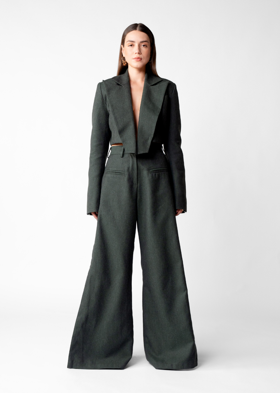 A woman stands against a light gray studio background wearing a dark charcoal gray tailored jumpsuit with wide legs, a structured collar, and a thin waist detail.