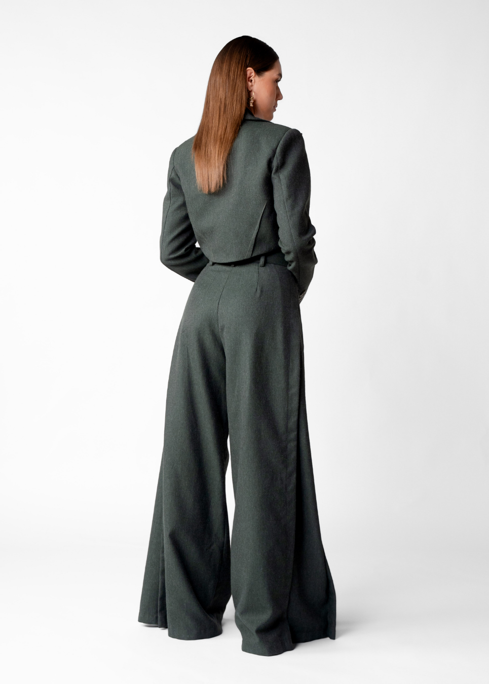 A woman shown from the back wearing a dark gray-green tailored jumpsuit with wide legs and a cropped jacket, photographed against a clean light gray studio background.