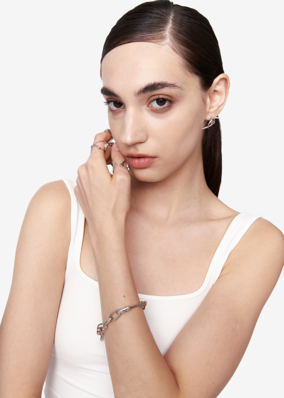 A model with dark hair in a ponytail wearing a white tank top, showcasing silver jewelry including a silver chain bracelet on her wrist and silver hoop earrings on her ear, against a plain white background.