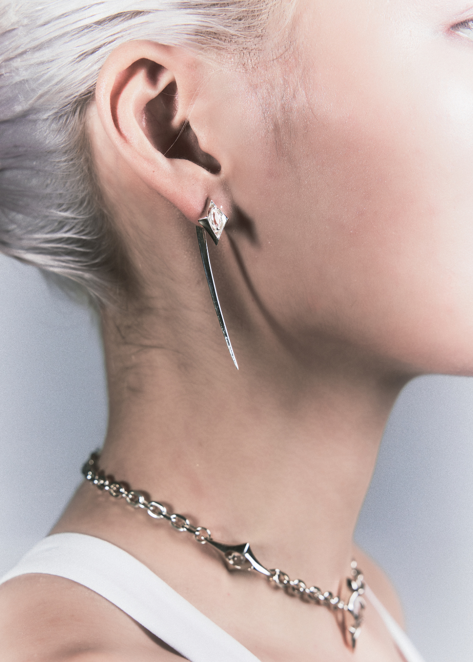 A close-up side profile of a model with short platinum blonde hair wearing a long, sharp silver dangle earring and a matching silver chain necklace.