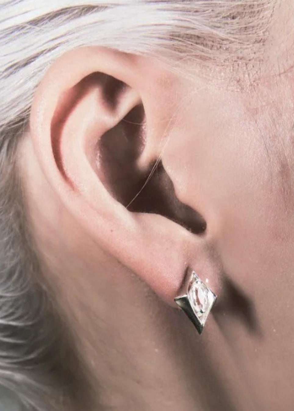 A close-up of a person's ear wearing a small silver geometric stud earring with a clear gemstone.