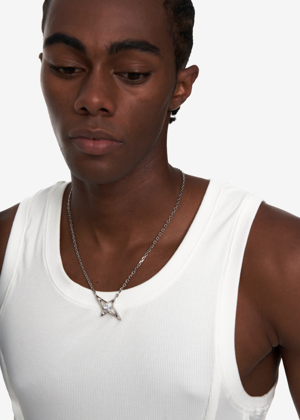 A close-up of a man wearing a silver chain necklace with a bow pendant, paired with a white ribbed tank top against a light gray background.