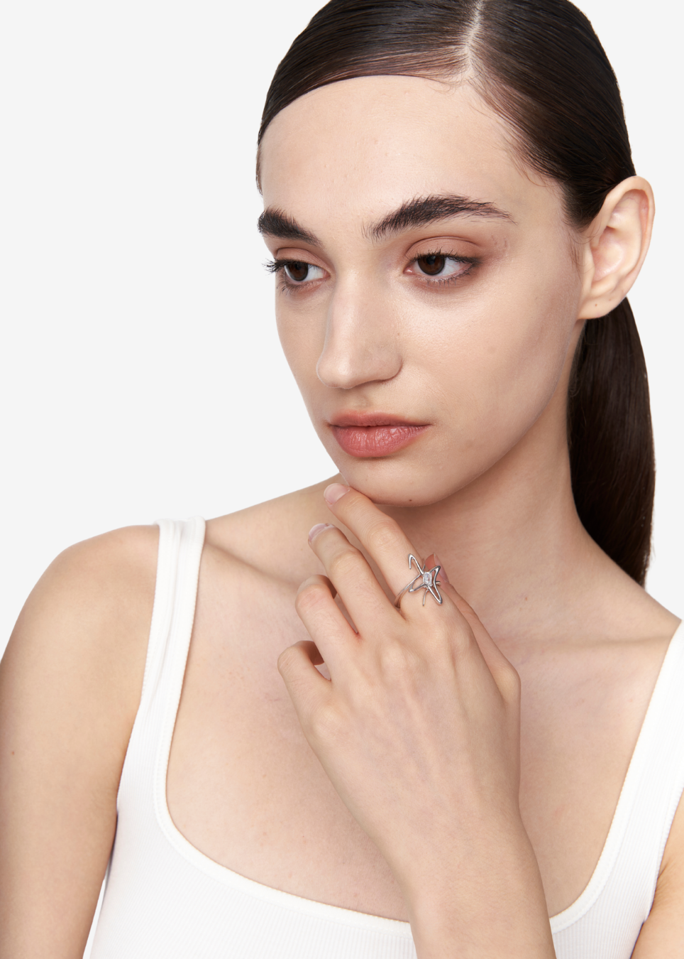 A close-up of a model wearing a silver ring with a star-shaped design on her finger, paired with a white tank top against a clean white background.