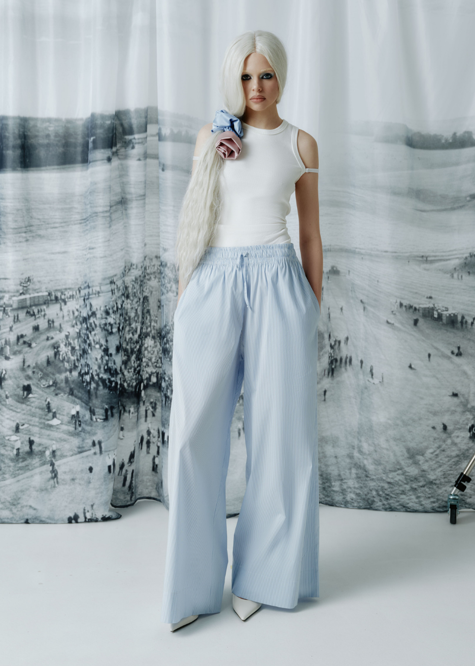 A model with long platinum blonde hair stands in front of a large black-and-white scenic backdrop, wearing a white sleeveless top and light blue wide-leg trousers.