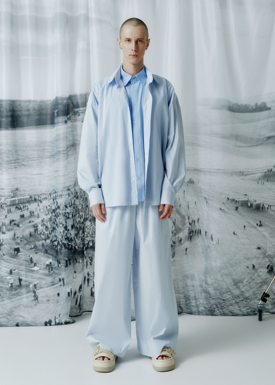 A model stands facing forward wearing a light blue oversized button-up shirt and matching wide-leg trousers, paired with beige slip-on shoes, in front of a large black-and-white landscape backdrop.