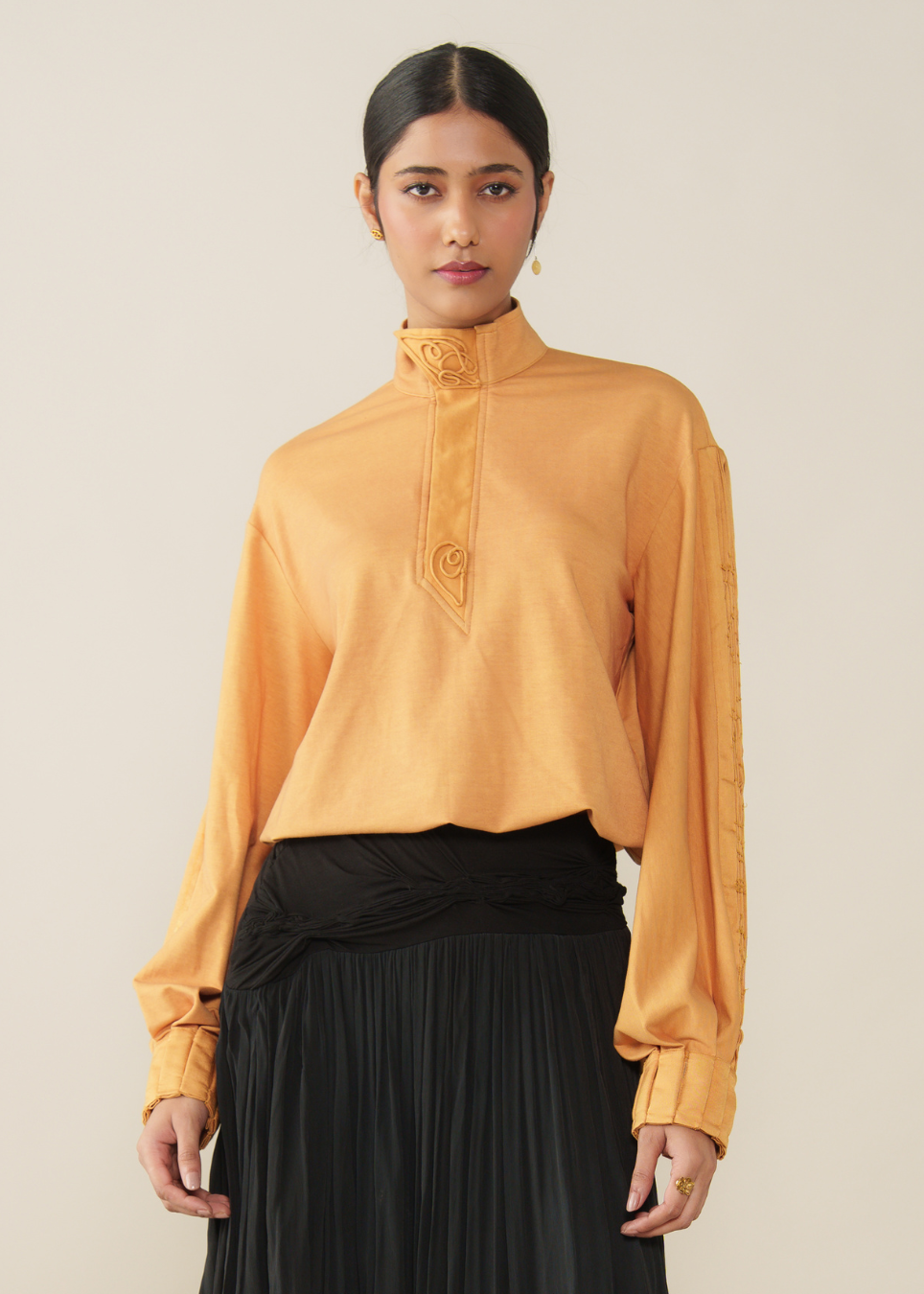 A woman modeling a mustard yellow long-sleeve blouse with a high mock neck and decorative embroidery on the chest, paired with a black pleated skirt, against a plain beige background.