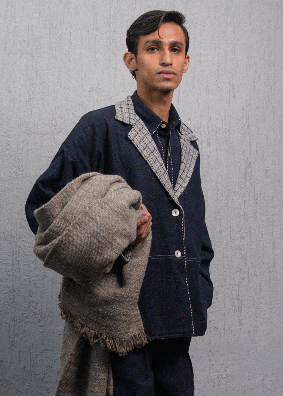 A man wearing a dark indigo jacket with a gray and white plaid collar, holding a folded gray woolen shawl against a textured gray background.