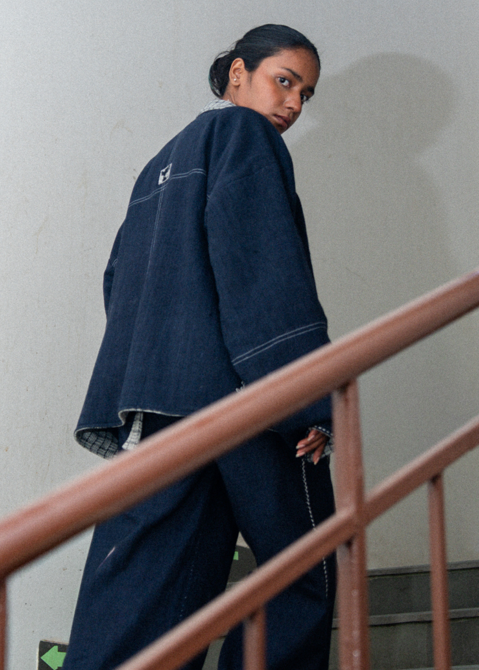 A model wearing an oversized dark indigo denim jacket and matching wide-leg jeans, captured from behind as they look over their shoulder while standing on a staircase with a wooden railing.