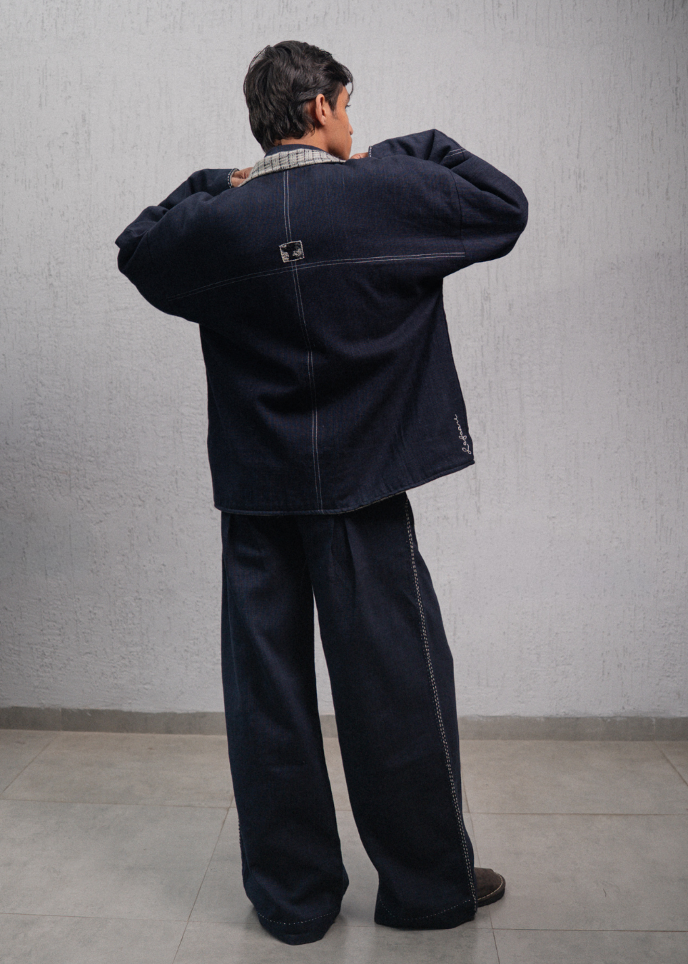 A person shown from the back wearing a dark indigo denim jacket and matching wide-leg denim pants, standing against a textured gray wall.