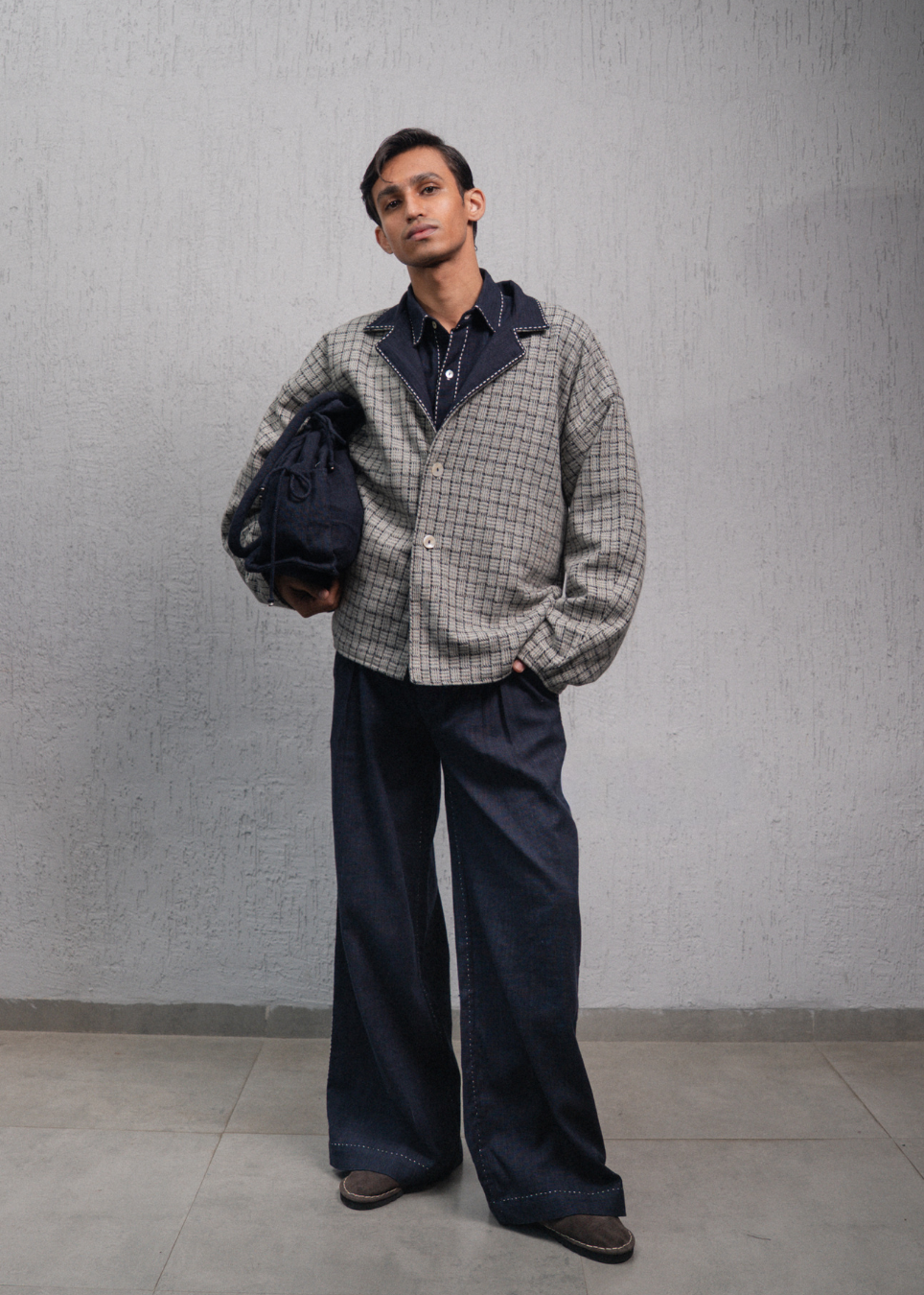 A man modeling a gray and black checkered overshirt with dark navy lining, paired with wide-leg dark blue jeans and holding a dark navy backpack against a textured gray wall.