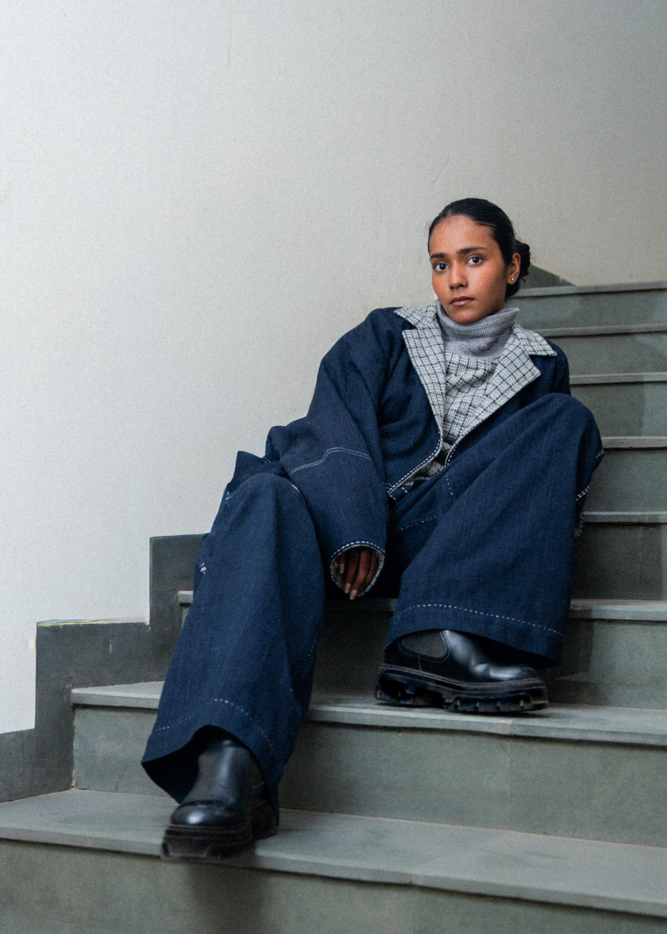 A model sits on concrete stairs wearing a dark blue oversized denim jacket and matching wide-leg jeans, layered over a gray patterned shirt and a light gray turtleneck, paired with black leather boots.