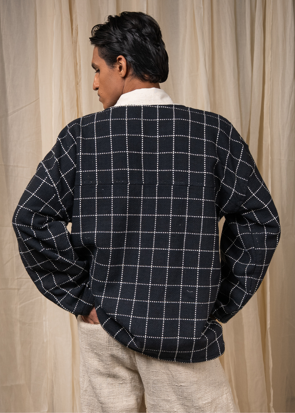 A man viewed from the back wearing a black long-sleeve jacket with a white dotted windowpane grid pattern, layered over a white collared shirt and paired with beige trousers, set against a neutral beige curtain backdrop.