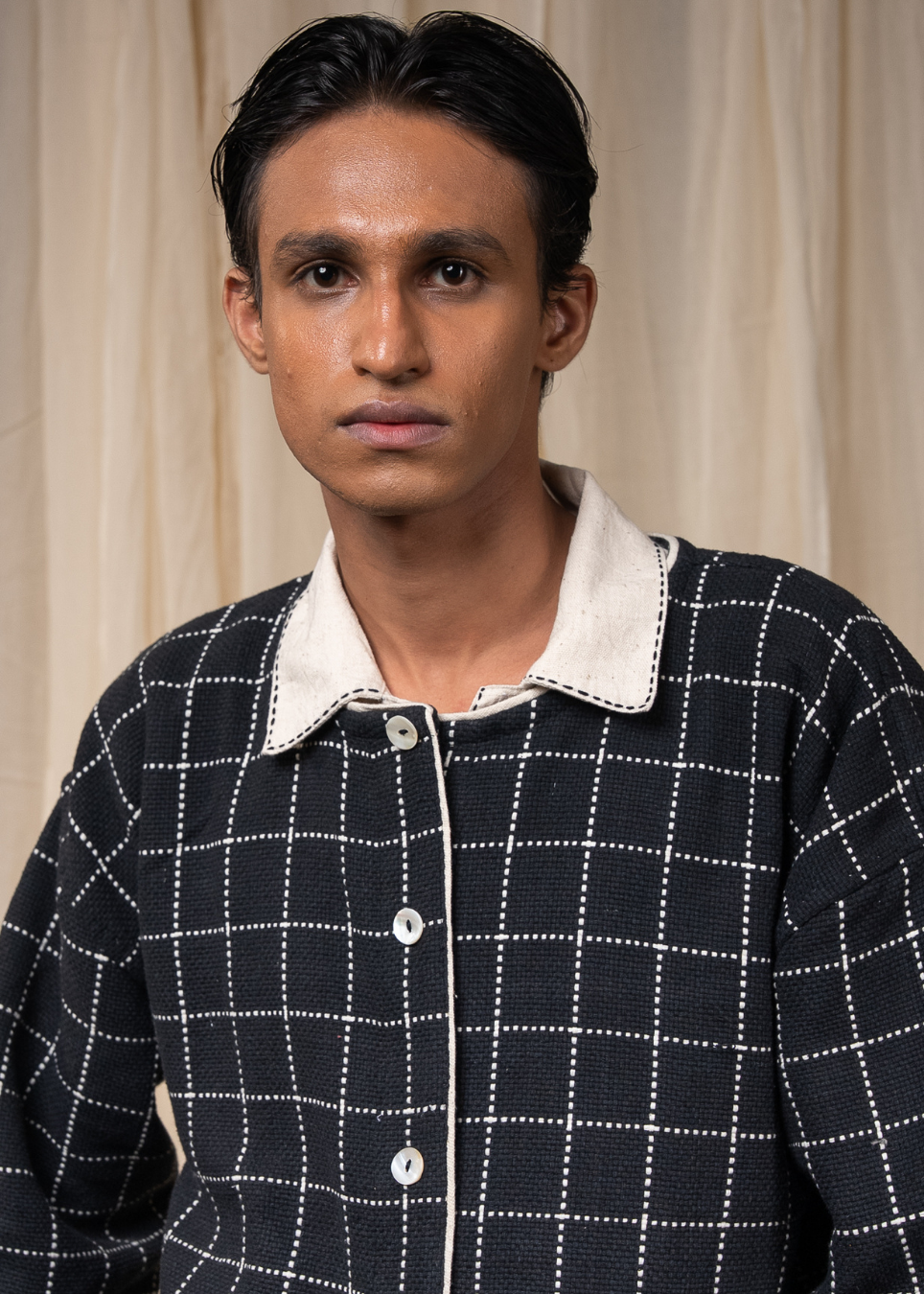 A man wearing a black knit button-up shirt jacket with a white grid pattern and a contrasting white collar, posed against a neutral beige curtain background.