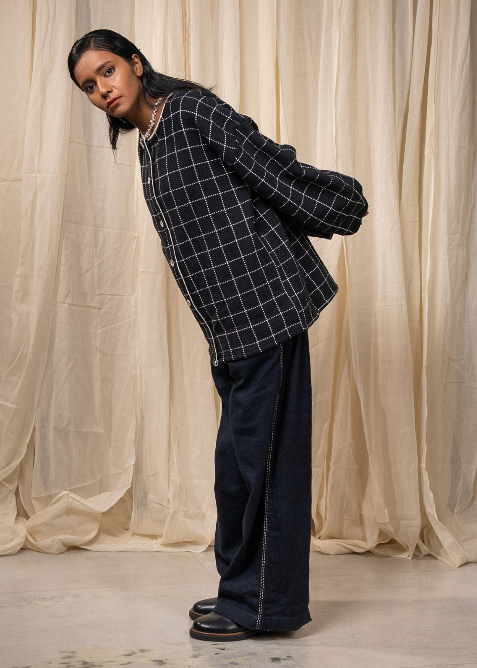 A model wearing a black and white windowpane plaid button-up shirt jacket and dark wide-leg jeans, standing in front of a draped beige fabric backdrop.