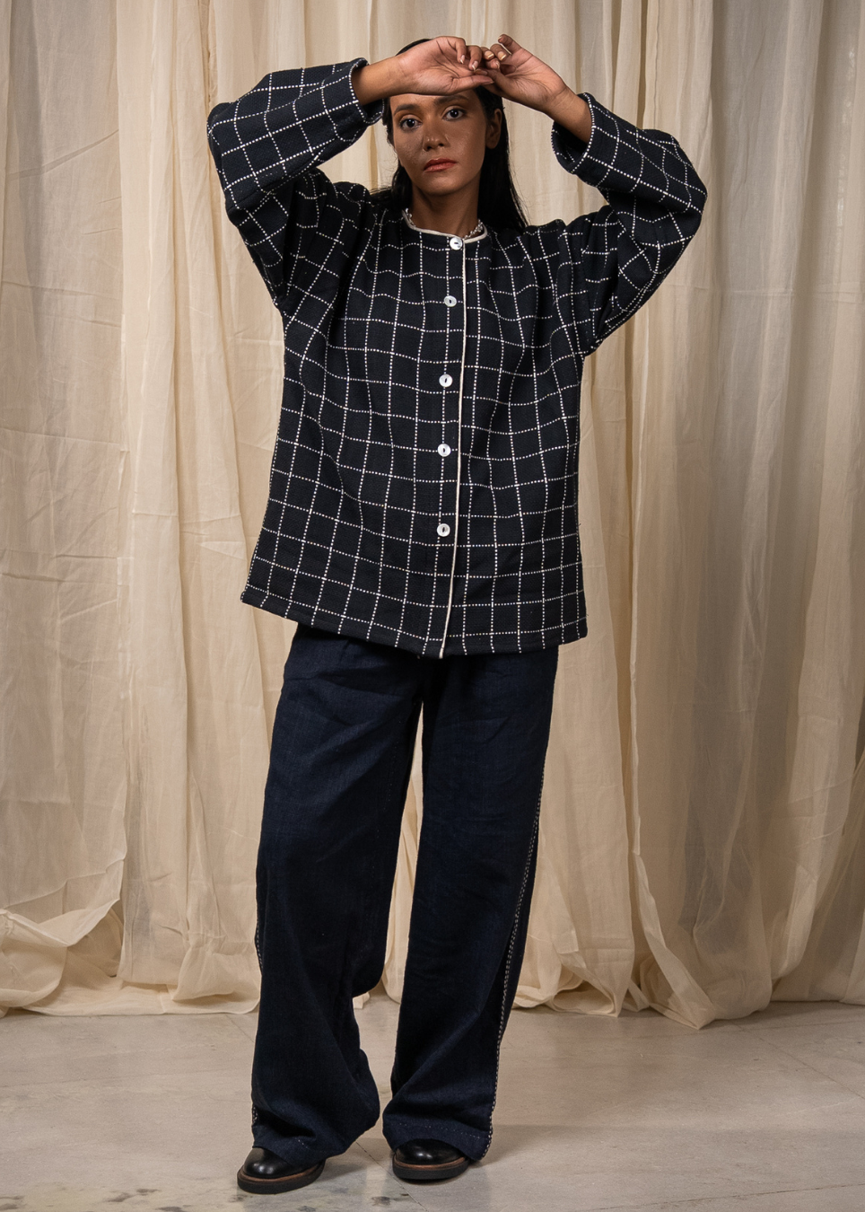 A woman modeling a dark navy blue jacket with a white windowpane plaid pattern, featuring a long-sleeve button-up top and matching wide-leg pants, posed in front of a draped beige fabric backdrop.