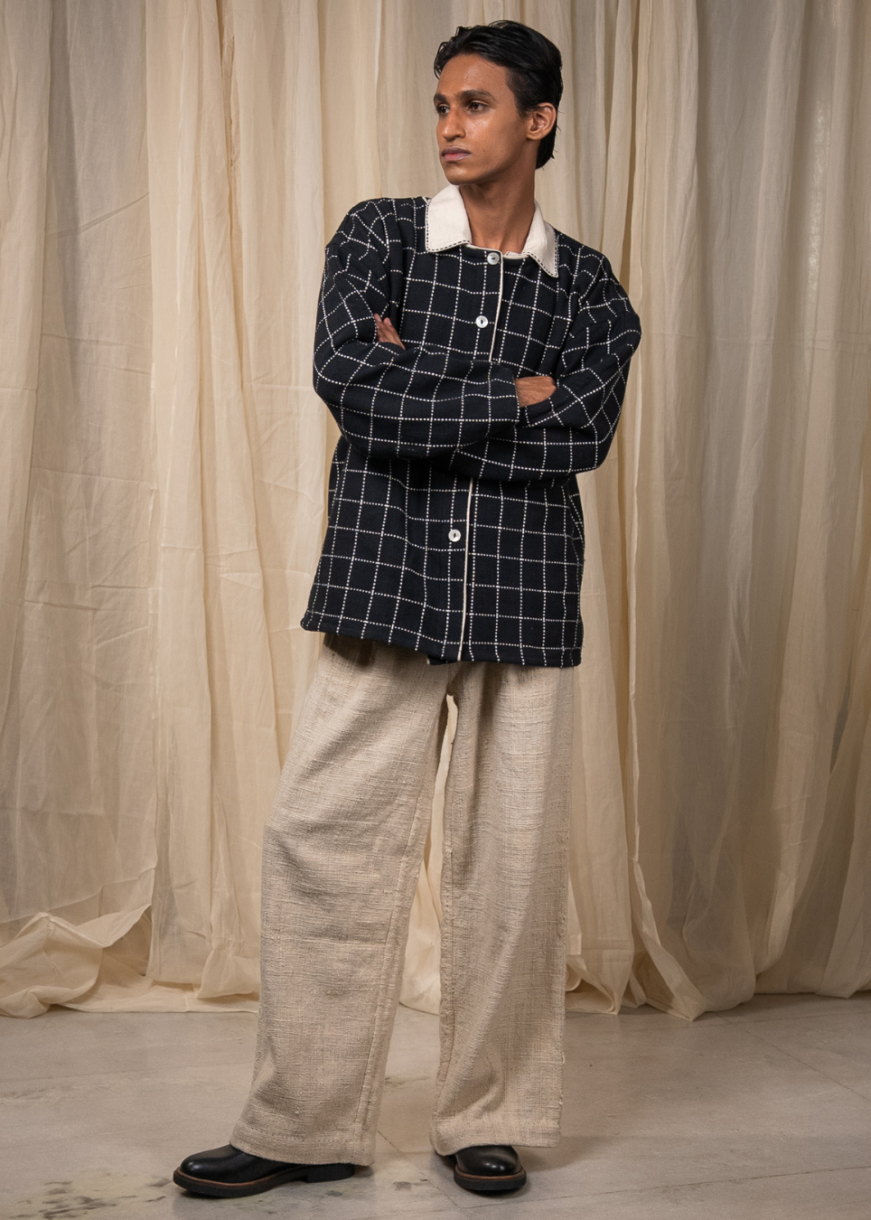 A man stands with arms crossed wearing a dark navy and white checkered button-up shirt jacket with a white collar, paired with loose beige linen pants and black shoes, set against a backdrop of draped beige fabric.