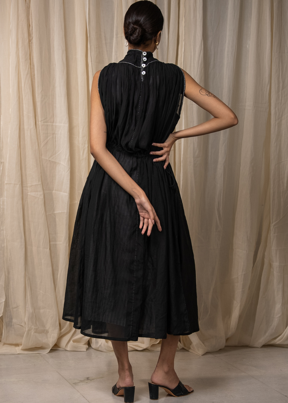 A woman shown from the back wearing a sleeveless black midi dress with a high collar and decorative silver buttons, standing in front of a beige curtain backdrop.