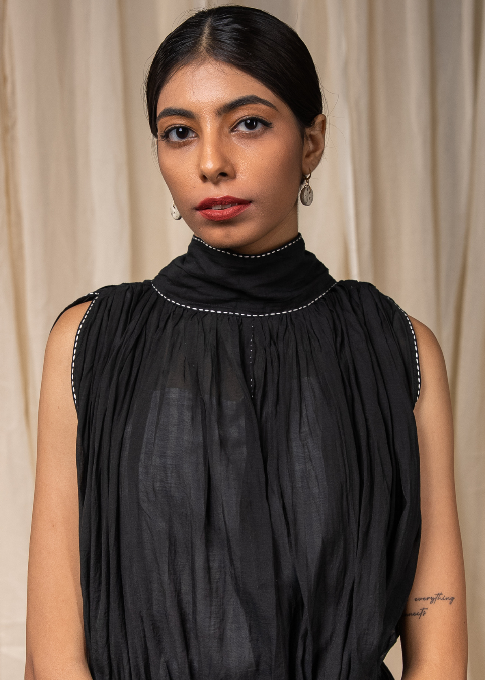 A woman wearing a sleeveless black high-neck pleated dress with subtle silver trim along the neckline and armholes, photographed against a soft beige curtain backdrop.
