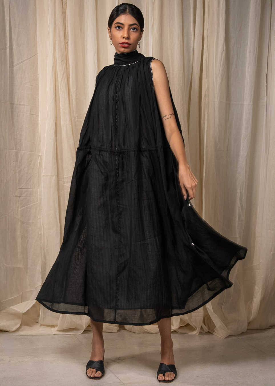 A woman models a sleeveless black midi dress with a high gathered neckline and a flowing, tiered skirt, standing against a draped beige fabric backdrop.