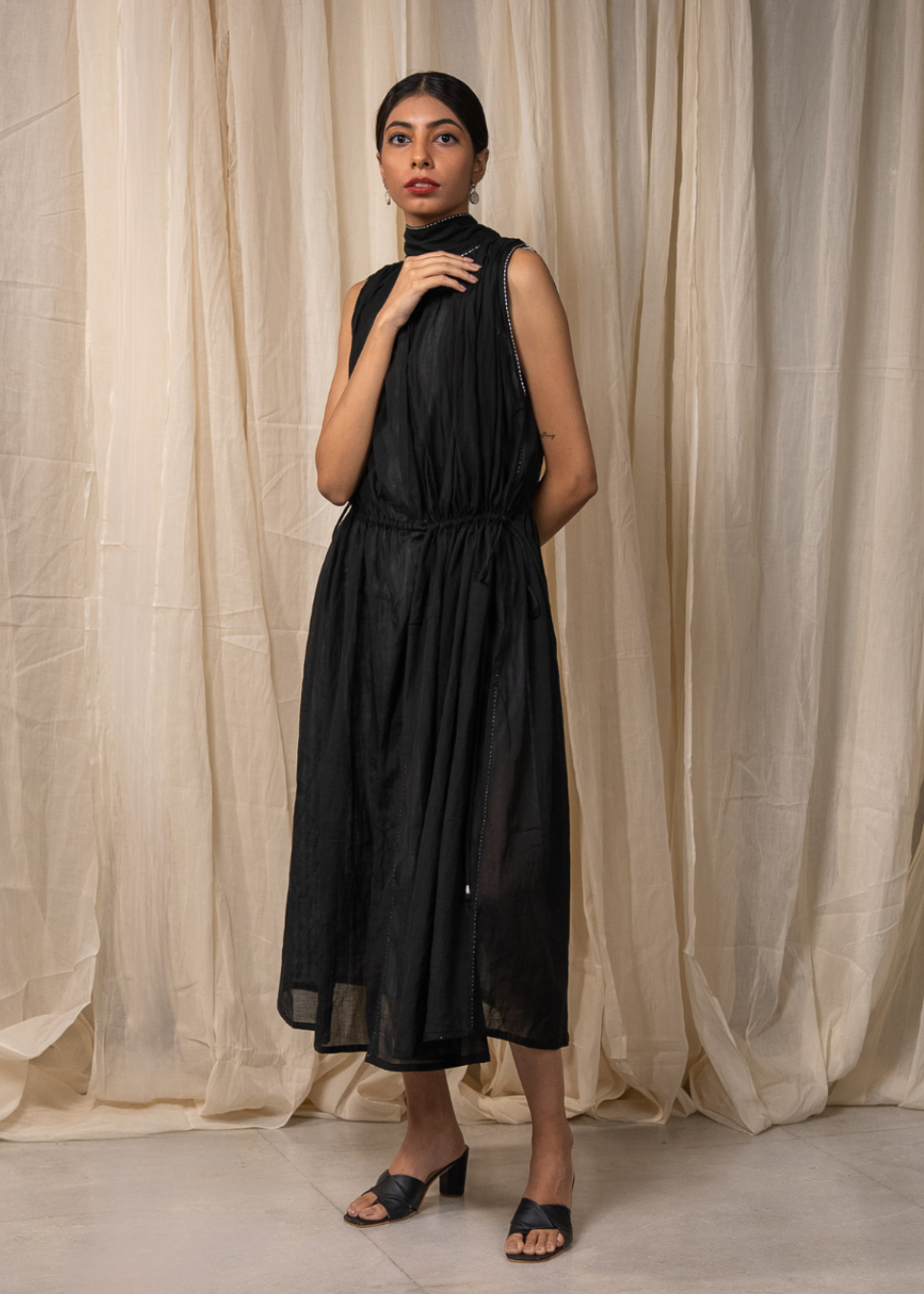 A woman modeling a sleeveless black midi dress with a high mock neck and drawstring waist, standing in front of draped beige fabric curtains.