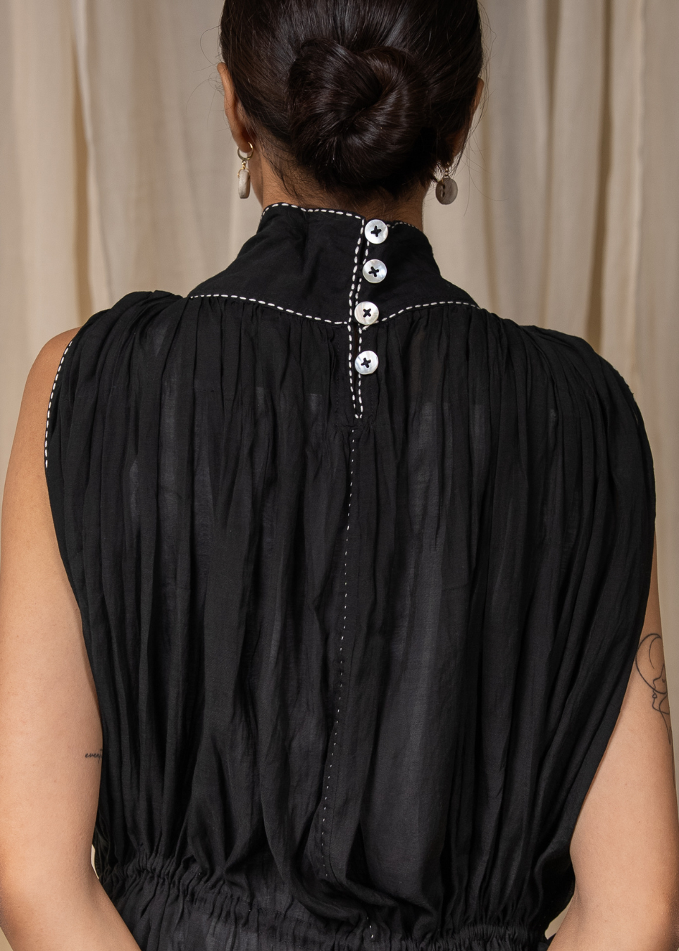 Back view of a woman wearing a sleeveless black dress with gathered pleated fabric, a high neckline, and three decorative silver buttons down the center back, set against a neutral beige curtain background.