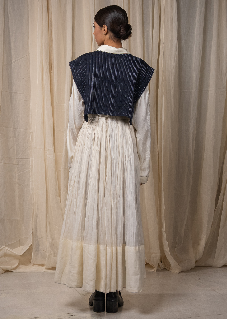 A model viewed from behind wearing a long-sleeved cream-colored pleated maxi skirt with a dark navy blue pleated cropped vest layered over the shoulders, standing in front of a draped beige fabric backdrop.