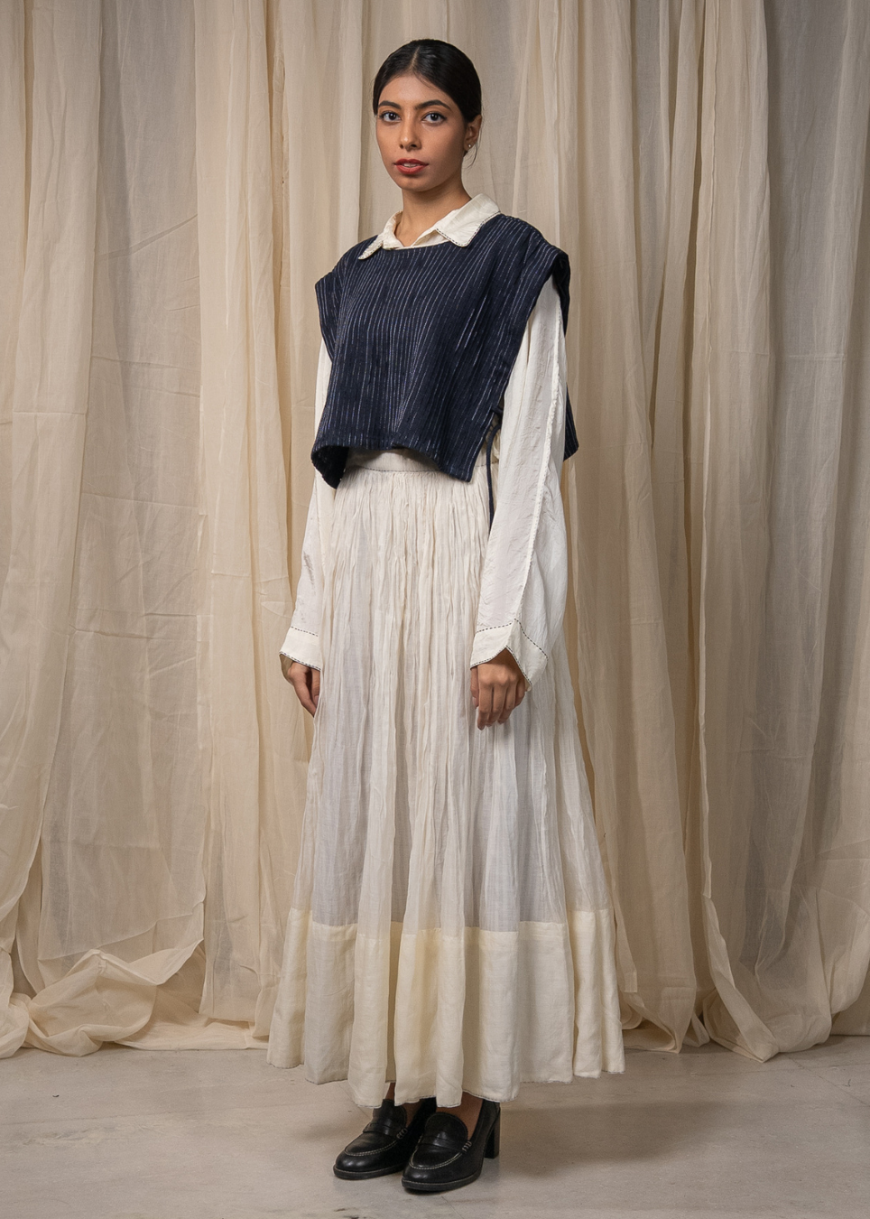 A model stands against a draped beige fabric backdrop wearing a long white pleated skirt, a white collared blouse, and a dark navy sleeveless pleated vest.