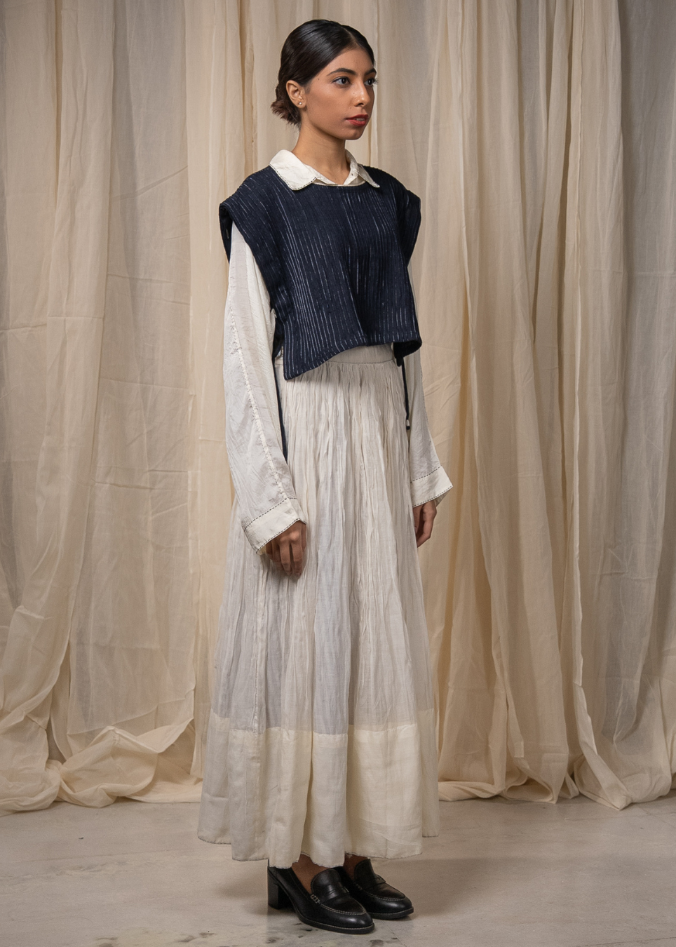 A model stands in front of draped cream fabric, wearing a long-sleeved white linen shirt with a dark navy blue pleated vest layered over the shoulders with a white maxi skirt, and black leather loafers.