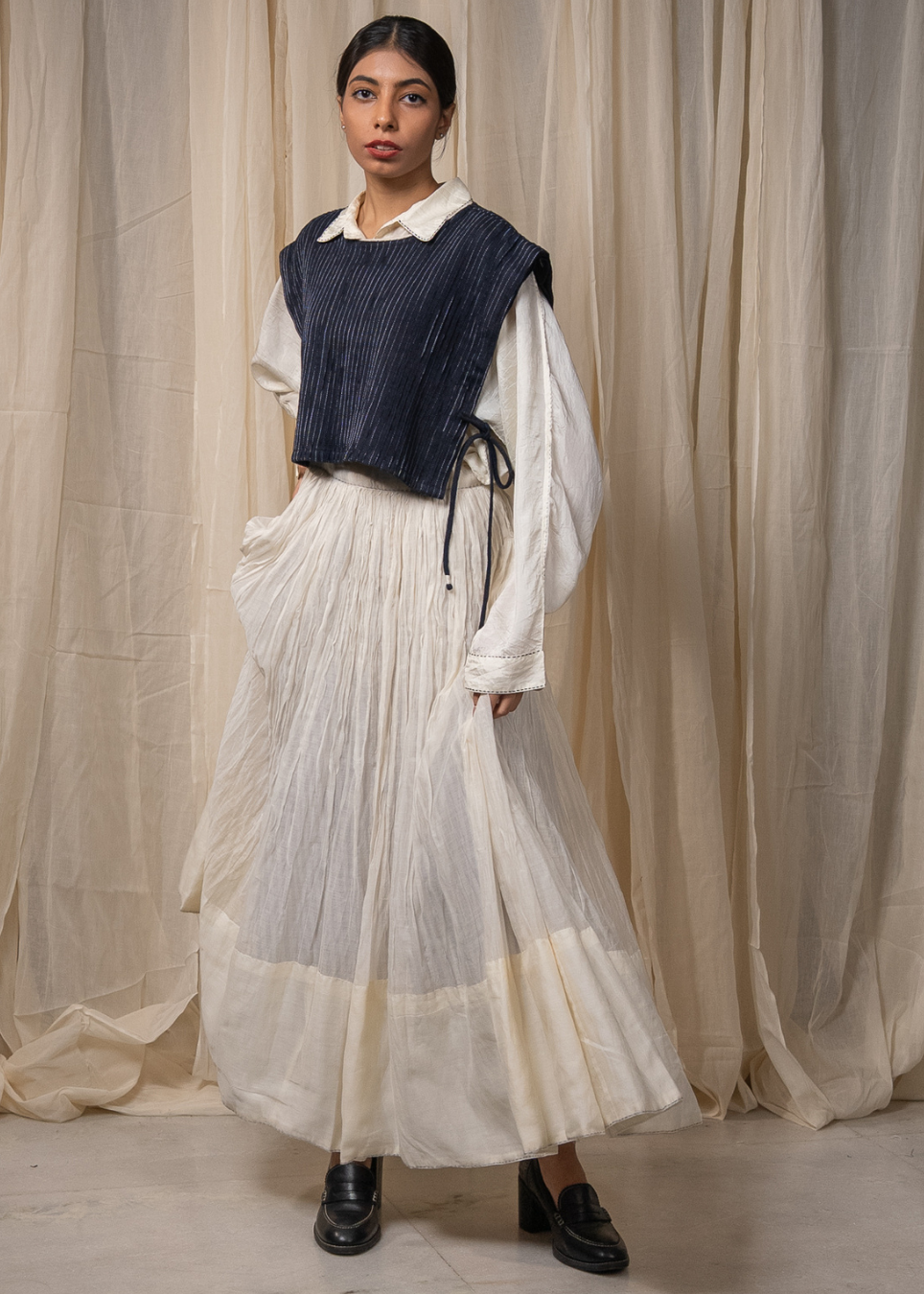 A model stands in front of draped beige fabric, wearing a long-sleeved white blouse under a dark navy pleated vest, paired with a long cream pleated skirt and black loafers.