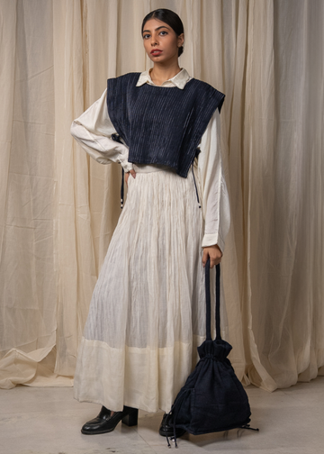 A model stands in front of beige draped curtains wearing a white collared shirt, a long pleated white skirt, and a navy blue ribbed vest. She is holding a matching navy blue drawstring bag.