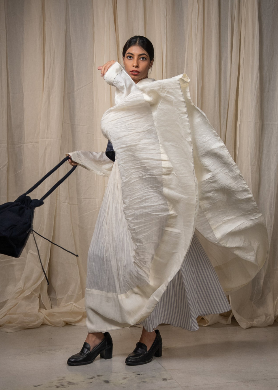 A model wearing a voluminous, textured off-white draped maxi skirt, holding a black bucket bag against a backdrop of beige fabric drapes.