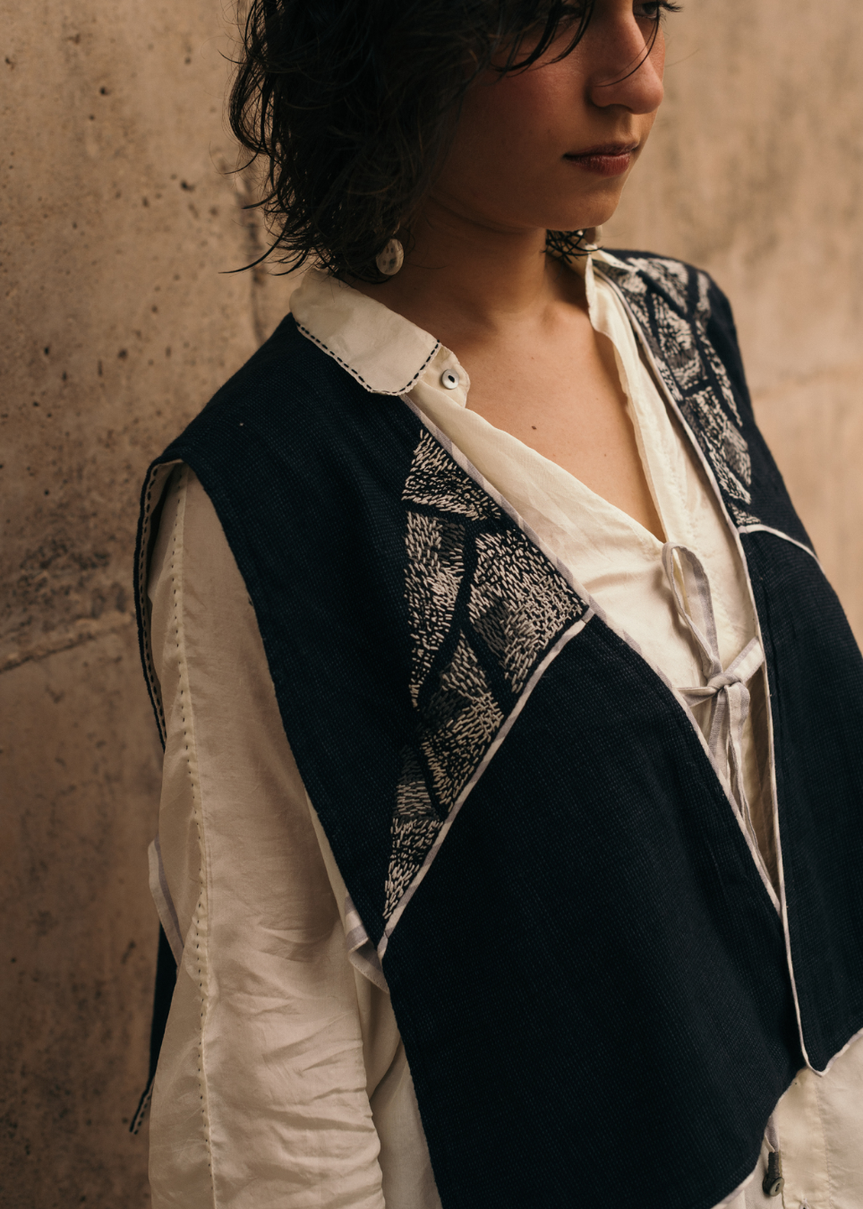 A person wearing a dark navy sleeveless vest with black-and-white patterned yoke panels over a white collared shirt, standing against a textured beige wall.