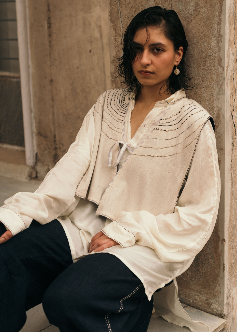 A woman wearing a cream-colored embroidered blouse and dark pants, sitting against a rustic stone wall.