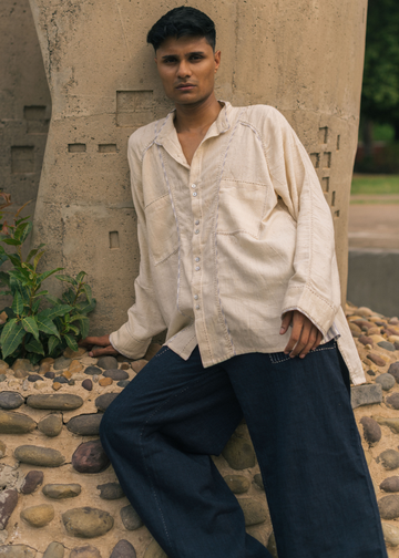 A man wearing a cream-colored linen button-up shirt and dark blue jeans, leaning against a textured concrete pillar in an outdoor setting.