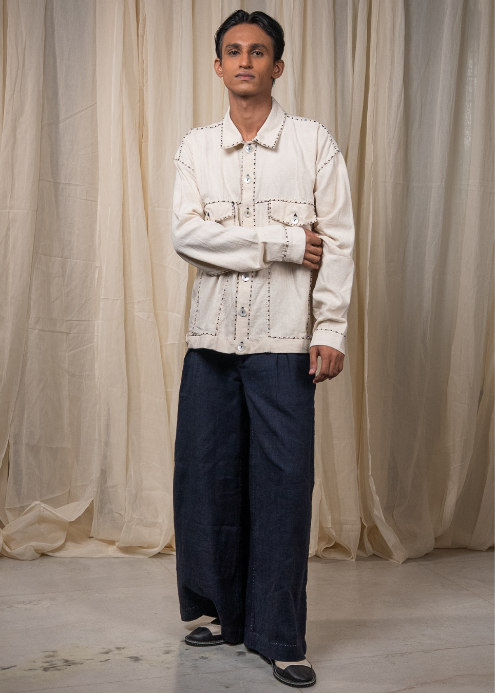 A man modeling a cream-colored, long-sleeve button-up shirt with decorative stitching and dark wide-leg trousers, standing in front of a draped beige fabric backdrop.